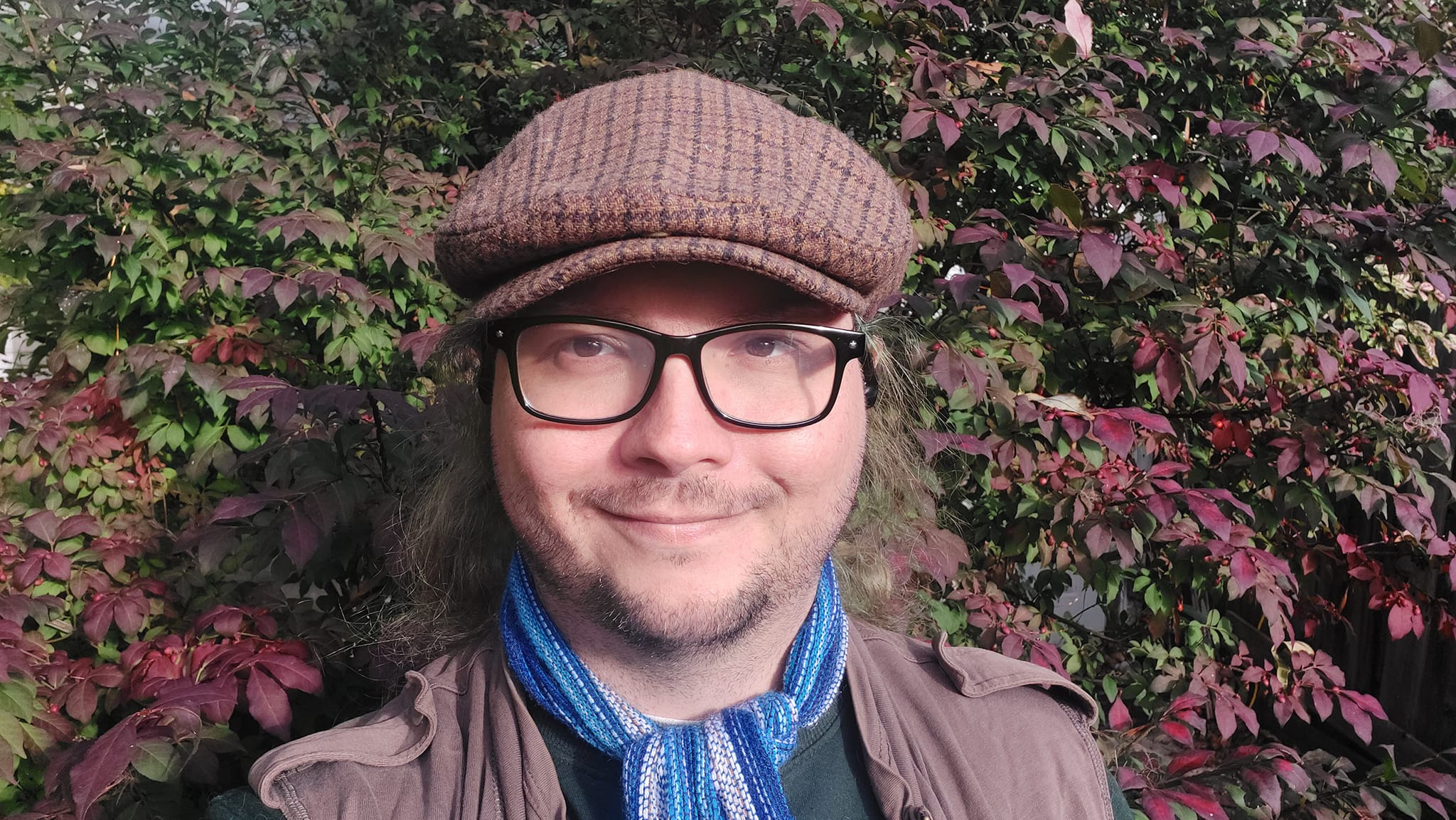 A man with long green hair wearing a green long sleave shirt with a brown zippered sweater vest, a blue scarf and a brown flat cap stand in front of a bush which has green to deep red leaves